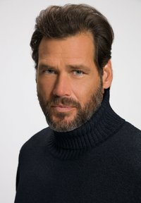 Dunkelblauer Rollkragenpullover mit strukturiertem gerippten Kragen, getragen von einem Mann mit kurzen, welligen Haaren und einem mittelgroßen Bart, vor einem schlichten Hintergrund.