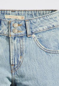 Jeans en denim bleu clair avec une taille très basse, dotés de deux boutons en métal, de coutures orange et d'une texture légèrement délavée. Taille 27.