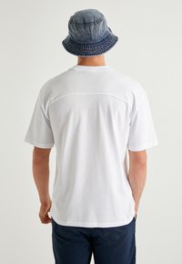 Camiseta de algodón blanca con mangas cortas, que presenta un detalle de costura en la parte posterior, combinada con un sombrero de cubo de mezclilla en tonos azules con un patrón texturizado.