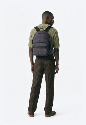 Bărbat purtând un pulover verde cu mâneci scurte și pantaloni maro cu dungi subțiri, cu un rucsac închis la culoare cu model, cu spatele spre privitor, pe un fundal alb.