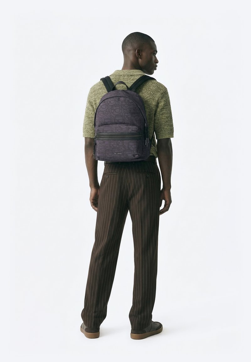 Hombre con un suéter verde de mangas cortas y pantalones marrones de rayas finas, con una mochila oscura con patrón, visto de espaldas sobre un fondo blanco.