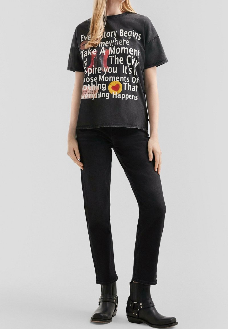 Femme portant un t-shirt imprimé noir surdimensionné, un jean noir et des bottines noires, debout devant un fond uni.