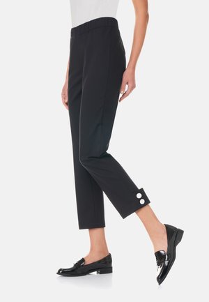 Nainen pukeutunut mustiin nilkkapitkiin housuihin, joissa on napit saumassa, ja mustiin kiiltonahkaisiin loafereihin, seisoo yhdellä jalalla.