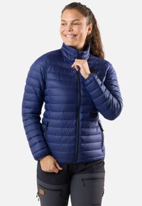 Laikka suojatakki, jossa on korkea kaulus, vetoketju, sivutaskut ja teksturoitu pinta. Yhdistetty tummiin housuihin neutraalilla taustalla.