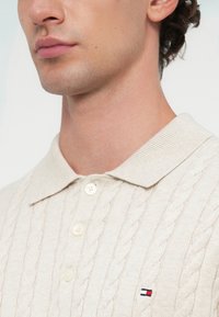 Beige Kabelstrick-Polohemd mit spitzem Kragen, ausgestattet mit drei weißen Knöpfen und einem kleinen Logo in Rot, Weiß und Blau auf der Brust.