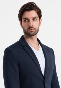 Marineblazer über einem weißen V-Ausschnitt-Hemd. Der Blazer verfügt über einen Reverskragen und Vordertaschen und zeigt eine strukturierte, aber glatte Textur.