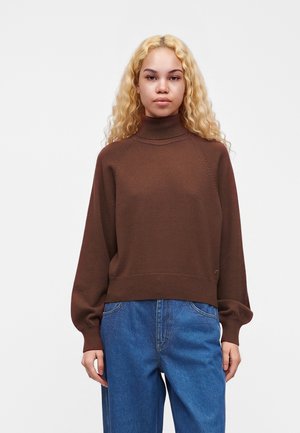 Giovane donna con lunghi capelli biondi e ricci, che indossa un maglione marrone a collo alto e jeans blu, in piedi contro uno sfondo bianco semplice.