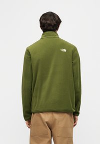 Άνδρας που φοράει πράσινο ελιά φλις μπουφάν με το λογότυπο The North Face στο πάνω μέρος της πλάτης, συνδυασμένο με καφέ παντελόνι, γυρισμένος με την πλάτη σε απλό φόντο.