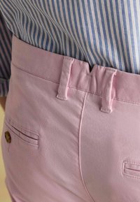 Jasnoróżowe bawełniane spodnie z płaskim przodem, dwoma bocznymi kieszeniami i guzikiem jako detalem. Połączone z niebiesko-białą koszulą w paski.