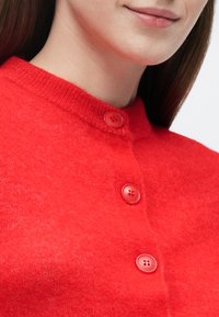 Czerwony kardigan z mieszanki wełny, z okrągłym kołnierzem, trzema fakturowanymi guzikami i miękkim, dzianym wykończeniem. Zbliżenie na detale i fakturę tkaniny.