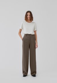 Woman with shoulder-length brown hair wearing a white short-sleeve shirt tucked into high-waisted wide brown pants and black shoes, standing.