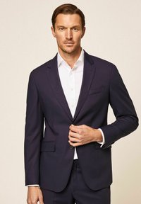 Mornarsko plava odijelo s jednim gumbom, oštrim reverima i dva prednja džepa, u kombinaciji s bijelom košuljom ispod.