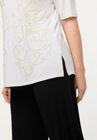 Top corto de manga corta blanco con intrincado bordado floral crema, aberturas laterales, combinado con pantalones anchos negros. Textura de tela suave.