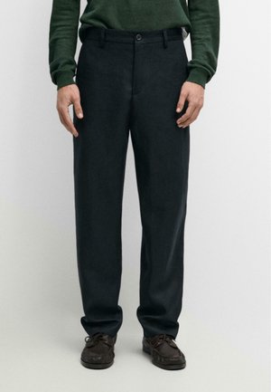 Mann trägt dunkelgraue maßgeschneiderte Hose, dunkelbraune Schnürschuhe und einen grünen Pullover mit langen Ärmeln, steht vor einem schlichten Hintergrund.