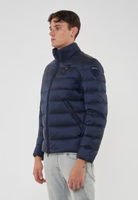 Giovane uomo con una giacca trapuntata blu navy con tasche zip e jeans chiari, in piedi di fronte a uno sfondo bianco semplice.