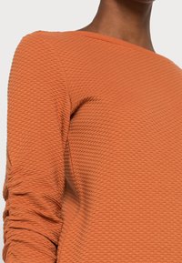 Oranssi neulepaita, jossa on hunajakenno-kuvio, pyöreä pääntie ja rypytetyt hihat. Huomioi materiaali ja suunnitteluelementit.