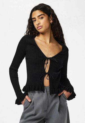 Pikka lokkis juustega naine kannab esiküljelt seotud musta soonikkoega kampsunit ja halli triibulist püksipukki, käed taskutes, valge taustaga.