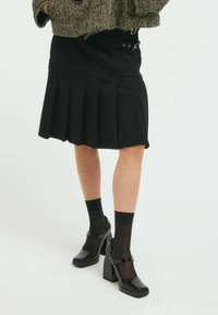 Fustă plisată neagră cu talie înaltă, asortat�ă cu pantofi Mary Jane negri lucioși și ciorapi negri transparenți, având o jachetă texturată în model herringbone.