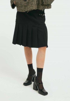 Schwarzer, plissierter Rock mit hoher Taille, kombiniert mit glänzenden, schwarzen Mary Jane-Schuhen und durchsichtigen, schwarzen Socken, dazu eine strukturierte Fischgrät-Jacke.