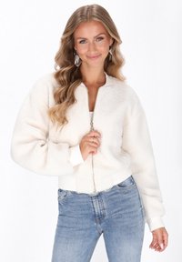 Giacca bomber bianca e morbida con zip frontale, polsini e orlo a coste. Indossata sopra una maglietta bianca e abbinata a jeans di denim blu chiaro.