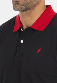 Homme portant un polo noir avec un col rouge et un logo de cheval brodé en rouge, avec des boutons blancs sur la patte de boutonnage.