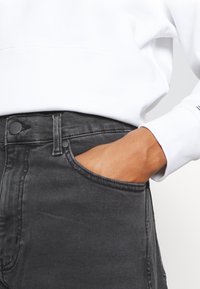 Jeans de mezclilla negra con corte recto, que cuentan con un bolsillo frontal y un cierre de botón. La tela tiene una ligera textura y un aspecto desgastado.