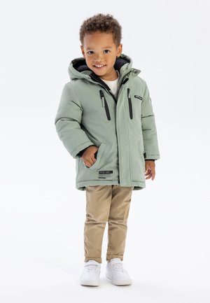 Mintgrüne, isolierte Jacke mit Kapuze, ausgestattet mit schwarzen Reißverschlüssen und Taschendetails, kombiniert mit beigen Hosen und weißen Sneakers.