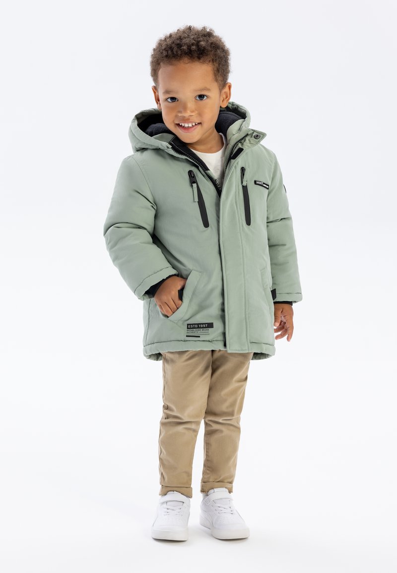Veste isolante vert menthe avec une capuche, dotée de zips noirs et de poches accentuées, associée à un pantalon beige et des baskets blanches.
