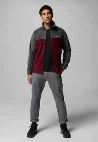 Giacca in pile rossa e grigia con zip frontale, pantaloni cargo grigi e scarponi neri da trekking; texture liscia e colori contrastanti.