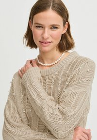 Un pull en tricot beige présente des motifs texturés, de grosses mailles en torsades et des perles décoratives le long des manches et du corps.