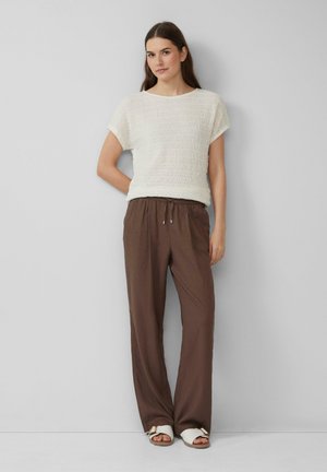 Crème textuur top met korte mouwen, gecombineerd met losse bruin broeken met een elastische tailleband en witte sandalen met een bandje.