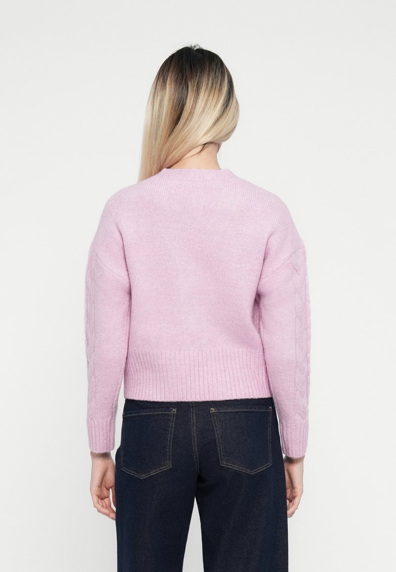 Pull en tricot rose avec un col rond, motif en torsades sur les manches et ourlet côtelé, associé à un jean bleu foncé.