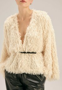 Kremowa kurtka z futra syntetycznego z głębokim dekoltem w literę V, oversize'owymi rękawami i czarnym paskiem w talii. Miękka faktura, casualowy design i brak zapięcia.