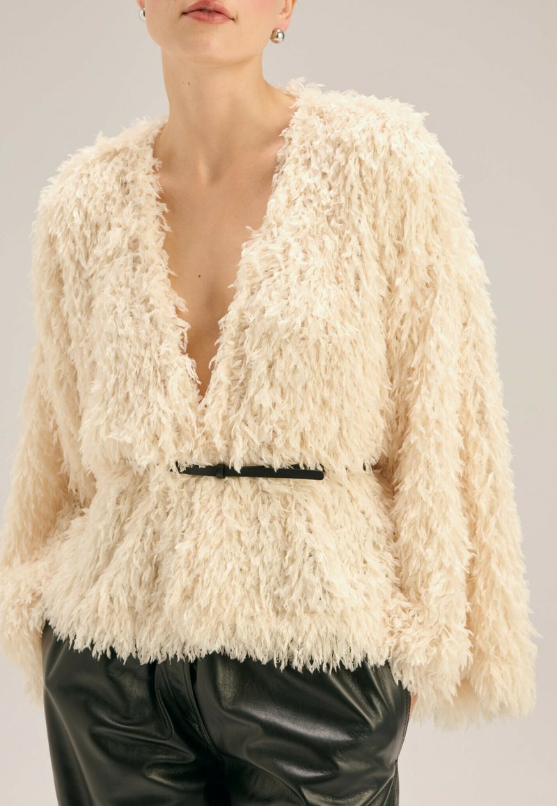 Kremowa kurtka z futra syntetycznego z głębokim dekoltem w literę V, oversize'owymi rękawami i czarnym paskiem w talii. Miękka faktura, casualowy design i brak zapięcia.