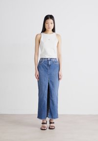 Hvit ermeløs topp med logo, kombinert med et langt denimbånd med splitt foran. Svarte sandaler fullfører antrekket.