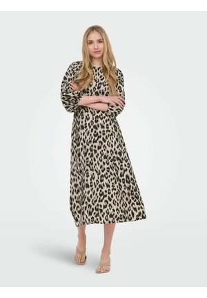 Femme aux longs cheveux blonds portant une longue robe à imprimé léopard et des sandales à talons beige, debout les bras croisés.