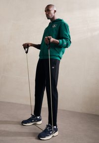 Zöld, zipzáras kapucnis pulóver logóval, fekete melegítőnadrág és sötétkék sportcipő. A modell edzőszalagokat tart a markolatnál, bemutatva a sportos öltözetet.