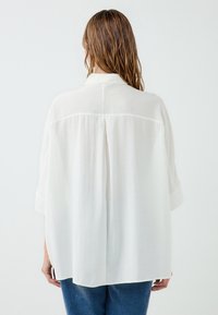 White oversized shirt made of lightweight fabric, featuring a classic collar, short sleeves, and a subtle back pleat detail.