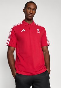 Punainen puuvillapaita, jossa on kolme valkoista raitaa kummallakin olkapäällä, Liverpool FC:n logo ja Adidas-logo rinnassa.
