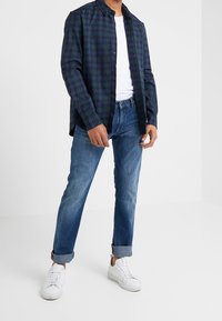 Geruite button-up shirt in marineblauw en groen; witte crew neck t-shirt; opgerolde denim jeans; witte sneakers met gestructureerde zolen.