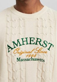 Krämfärgad stickad tröja med flätmönster. Har broderad text: "AMHERST," "Original sedan 1998" och "Massachusetts" i grönt och orange.