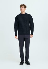 Pull en tricot avec un motif de câble bleu marine et un col rond, présentant une texture en grosse laine. Associé à un pantalon sombre et des chaussures noires.