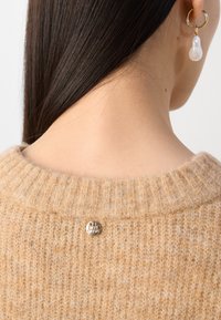 Pulover bej, tricotat cu model cu nervuri, cu guler rotund, având un detaliu cu un buton argintiu la baza gâtului. Textura moale este vizibilă.