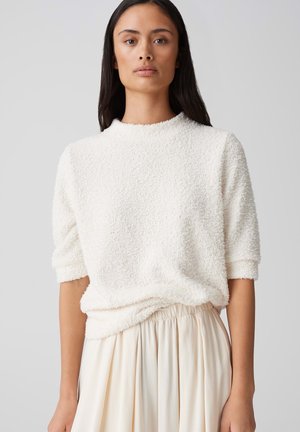 Weißer, strukturierter Pullover mit rundem Halsausschnitt und kurzen Ärmeln, kombiniert mit einem fließenden cremefarbenen Rock. Der weiche Stoff hat ein gemütliches Aussehen.