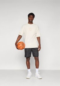 Gaiši bēša oversized T-krekls ar smalku grafiku, kopā ar melniem sieta šortiem un baltām sporta kurpēm, tur oranžu basketbola bumbu.