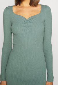 Robe à manches longues côtelée en vert pâle, présentant un décolleté carré et un détail de fronces au centre, fabriquée dans un tissu extensible.
