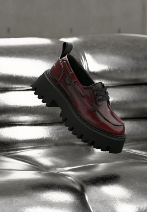 Dark red leather shoe with thick black lug sole floating above a shiny silver tufted surface, showing side and top details.