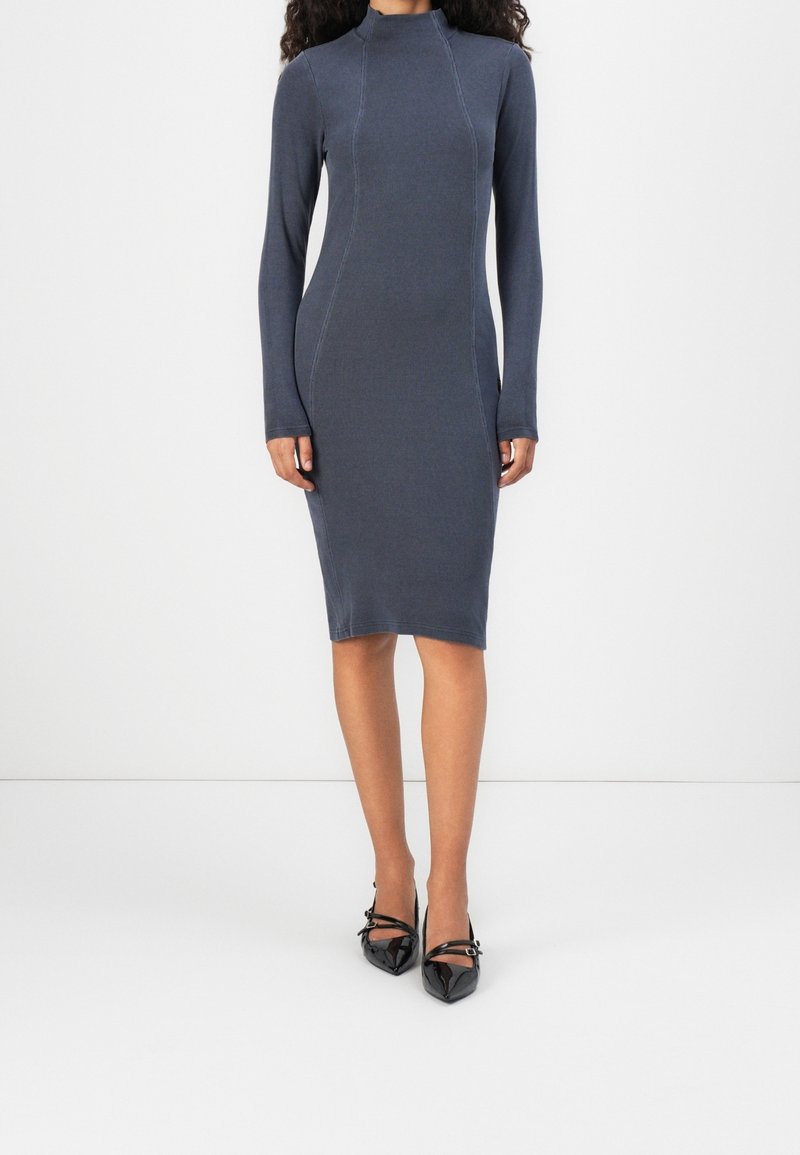 Femme portant une robe grise foncée ajustée, à manches longues, longueur genou, col montant, avec des escarpins noirs pointus à brides.