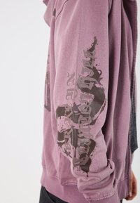 Μωβ φούτερ με σκούρα γραφικά εκτύπωσης στα μανίκια, κατασκευασμένο από μαλακό ύφασμα με άνετη εφαρμογή και ριμπ μανσέτες.