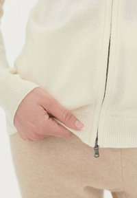 Krämfärgad zip-up tröja med ribbade ärmslut, texturerat tyg och detaljer i metall på dragkedjan. En hand greppar dragkedjan vid den nedre fållen.